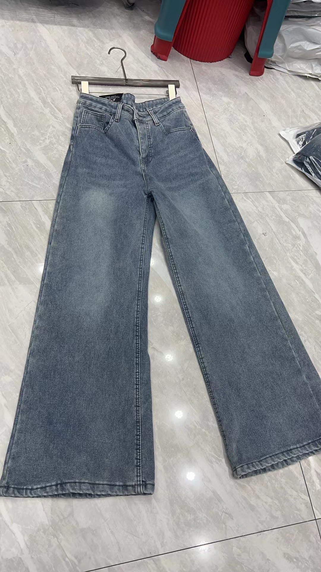 这是一张展示服装套图案例的白底3D图，呈现的是一条复古水洗浅蓝色的阔腿牛仔裤，挂在带有金属挂钩的木质衣架上，平铺于带有反光亮点的浅灰色大理石纹理地面。这条牛仔裤版型宽松，裤腿垂感自然，整体呈现出干净利落的服装展示效果，属于文档中介绍的服装批量生图服务里的白底3D图类产出案例。