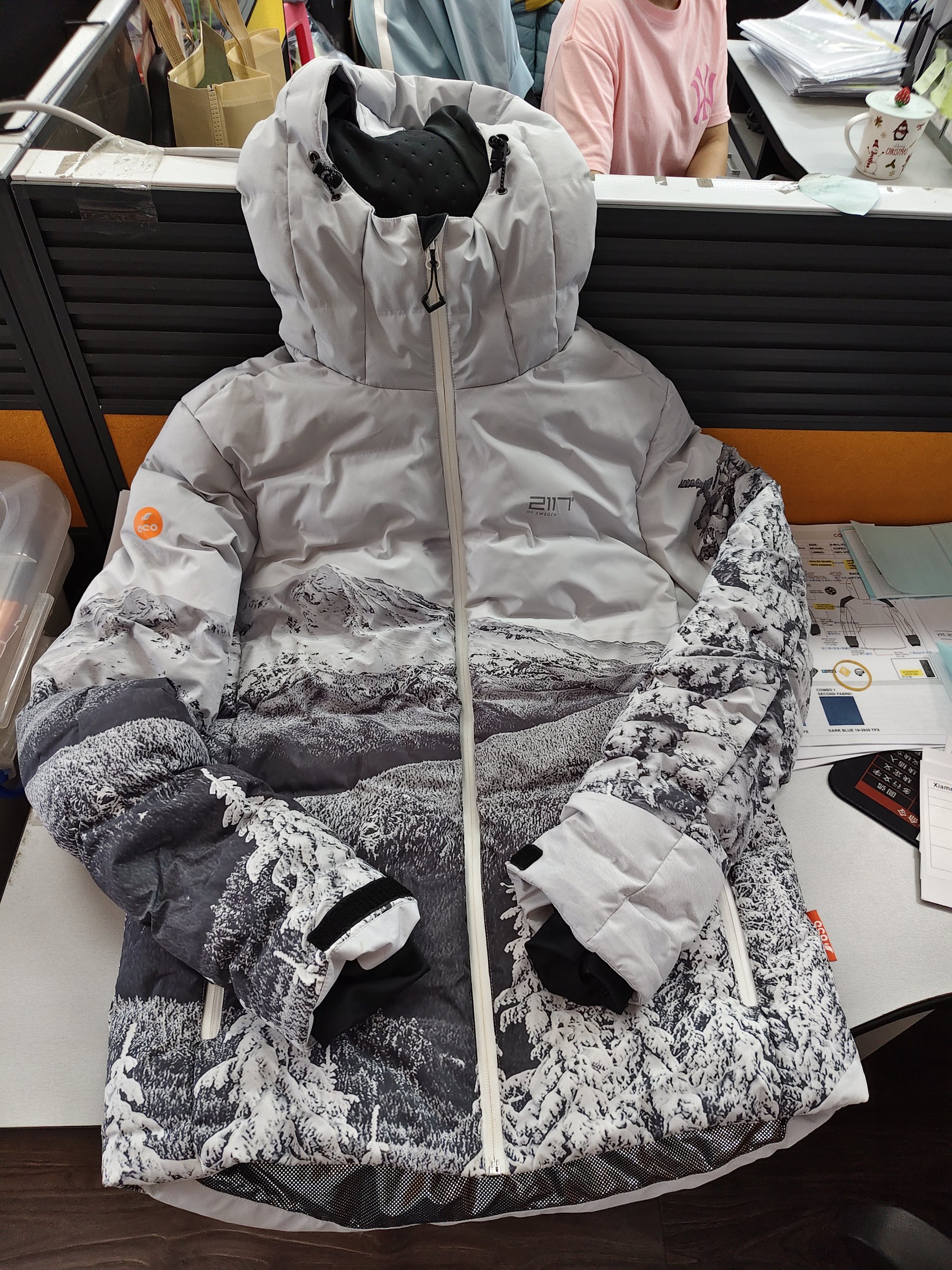 这是一件用于服装套图制作的白底3D示例服装，为连帽款式的滑雪服，整体采用灰、黑、白为主的雪山森林印花设计，兜帽处带有黑色护脸结构，正面配有白色拉链，左侧袖身带有橙色标识，袖口处可见黑白拼接细节。这件服装是前文批量生图功能中，用于生成白底3D图类型的服装案例，以直观展示该类批量生成的服装图所呈现的实际产品效果。