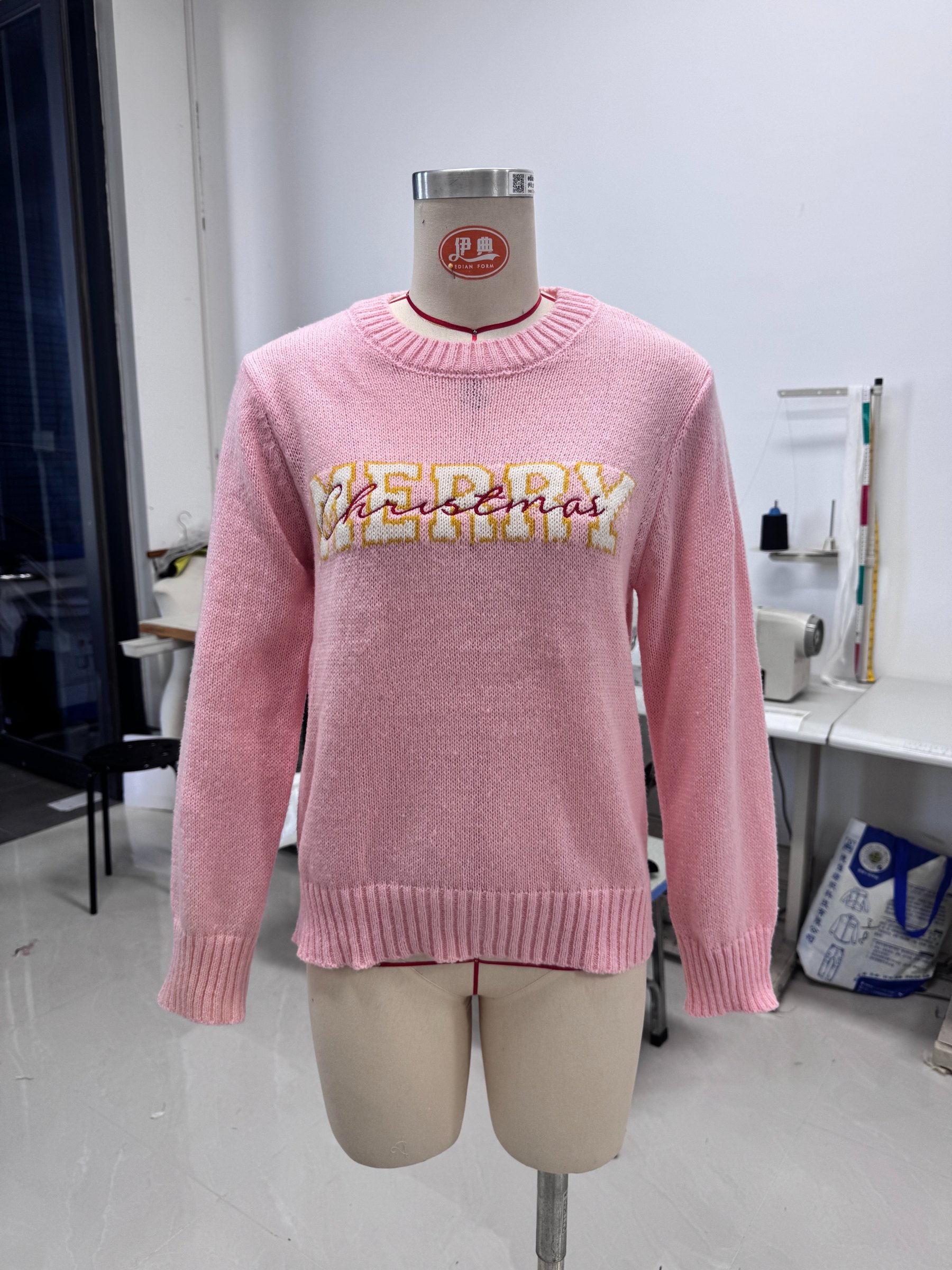 这张图片展示的是一件陈列在人体模型上的粉色圆领针织毛衣，毛衣胸前带有黄白配色的“MERRY Christmas”字母装饰图案。该图片对应的是批量生图案例中的白底3D图产出示例，体现批量生成服装相关套图里白底3D图的成果，用于直观展示该类服装的3D呈现效果，符合文档中关于服装套图案例的内容，是批量生成白底3D图的实际效果展示。