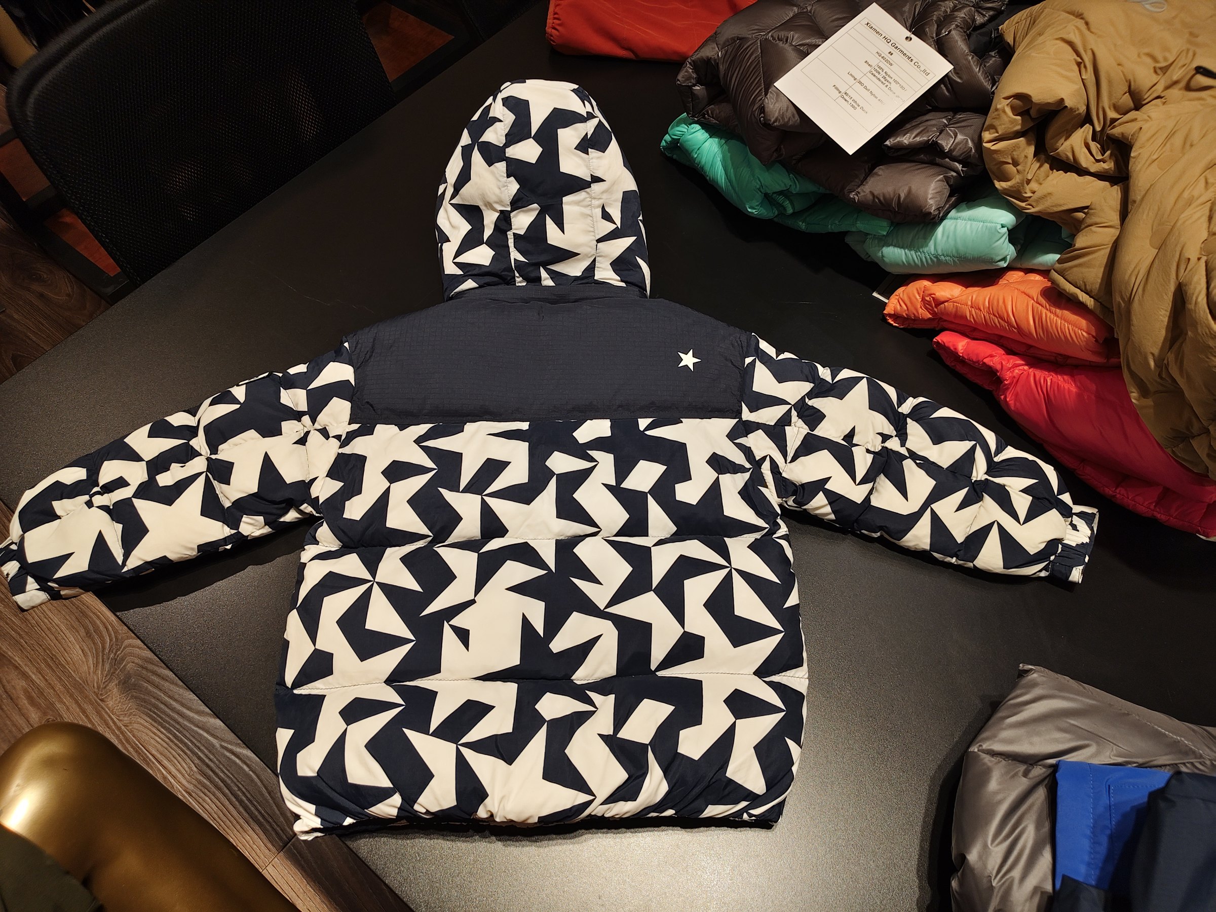 图片展示了一件服装，为黑色和白色几何图案的外套。外套背面有白色星星装饰，内里为黑色。外套平铺在深色桌面上，周围摆放着其他不同颜色和款式的服装，如红色、绿色、橙色、黄色等羽绒服。图片位于介绍服装套图相关内容中，可能是用于展示服装款式、图案及整体外观，辅助说明服装套图的样式特点。