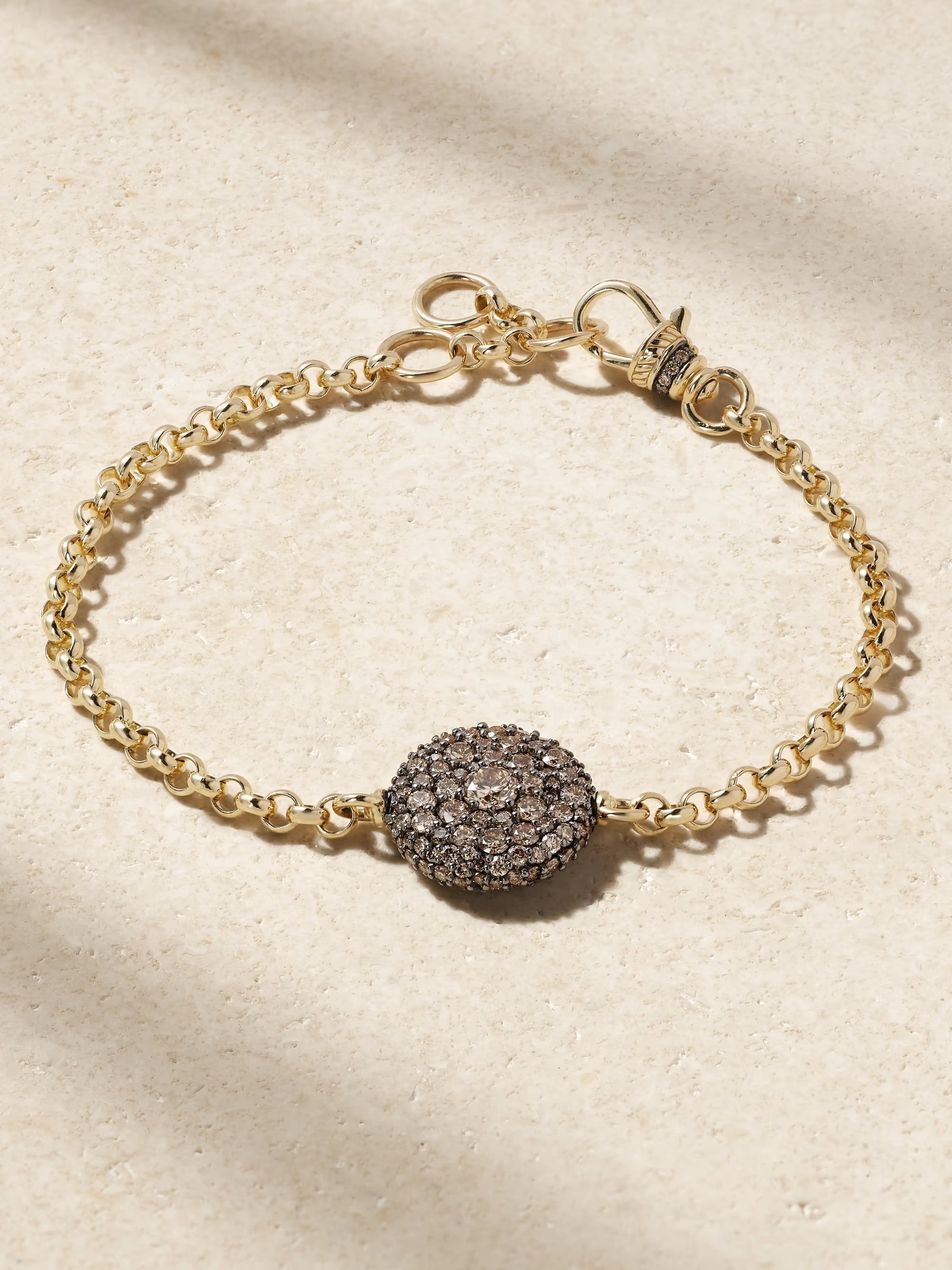 图片展示的是经典链节式手链（Rolo Chain Bracelet）。手链主体由金色链节组成，呈现出精致的链条质感。手链中间有一个圆形装饰，由多个小圆环组成，表面镶嵌着黑色和金色的装饰物，显得华丽且有层次感。手链两端各有一个金色的扣环，设计简洁实用。背景为浅色纹理表面，手链被放置在中央，整体呈现出优雅且时尚的外观。