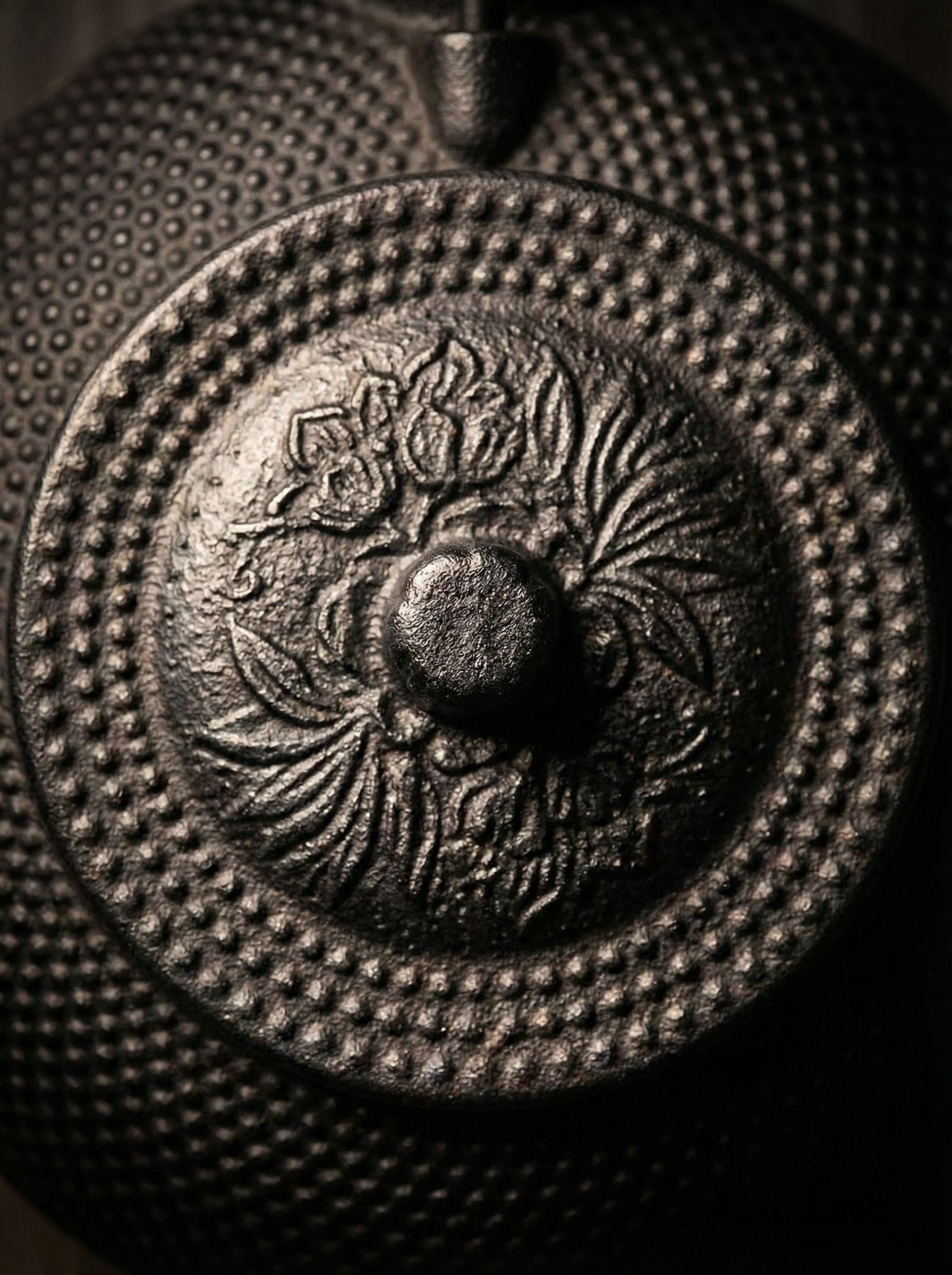 图片展示的是一个铸铁壶的壶盖特写。壶盖表面有密集的凸点装饰，中央有一个圆形凸起，周围环绕着复杂的花卉图案，包括花朵和叶子。壶盖整体部分呈圆形，边缘有明显的凸起。整体色调为深色，呈现出铸铁材质的质感。该图片可能用于展示商品的材质和工艺细节，与文档中商品套图效果展示的内容相关。