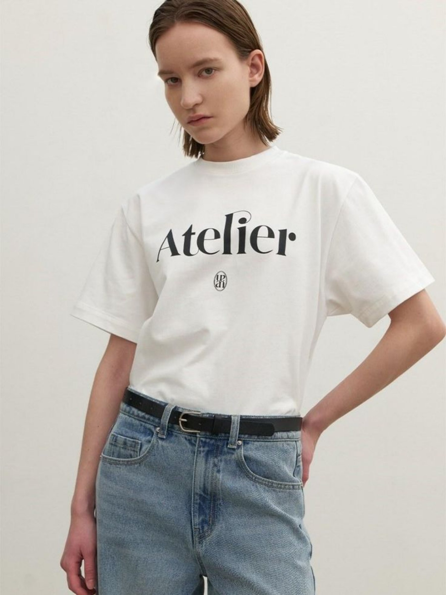 图片展示了一位穿着白色T恤的女性。T恤正面印有“Atelier”字样及“MG”标志，她搭配浅蓝色牛仔裤，腰间系着黑色皮带，双手插兜，背景为纯白色。该图片对应文档中“修改商品款式/形状”部分的示例，提示词为“将上衣改为V领短袖款式，保留上衣原有的橙色”，用于展示智能修图在修改商品款式方面的应用效果。